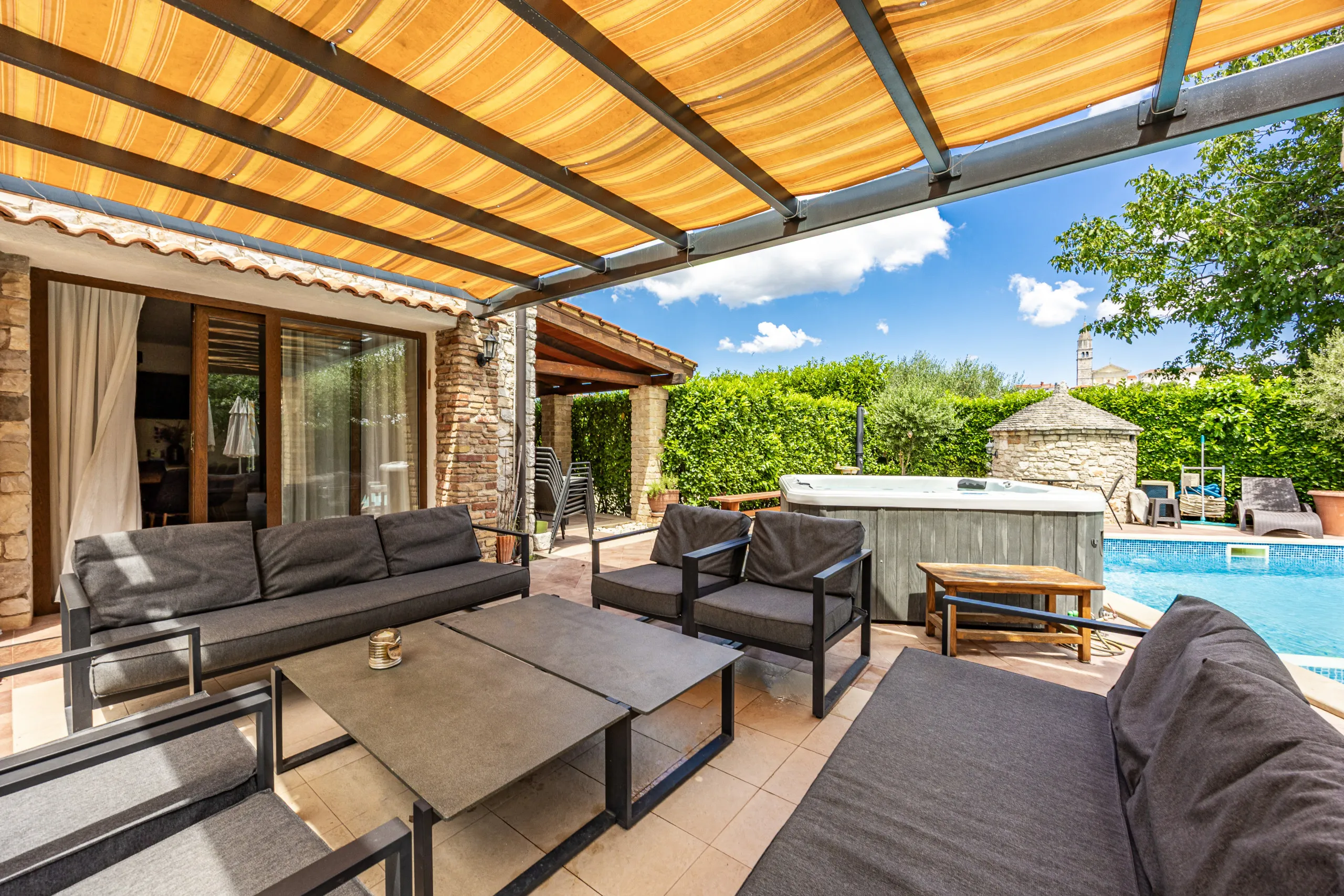 Sofas und niedrige Tische unter der gestreiften Pergolamarkise, daneben Whirlpool und Pool