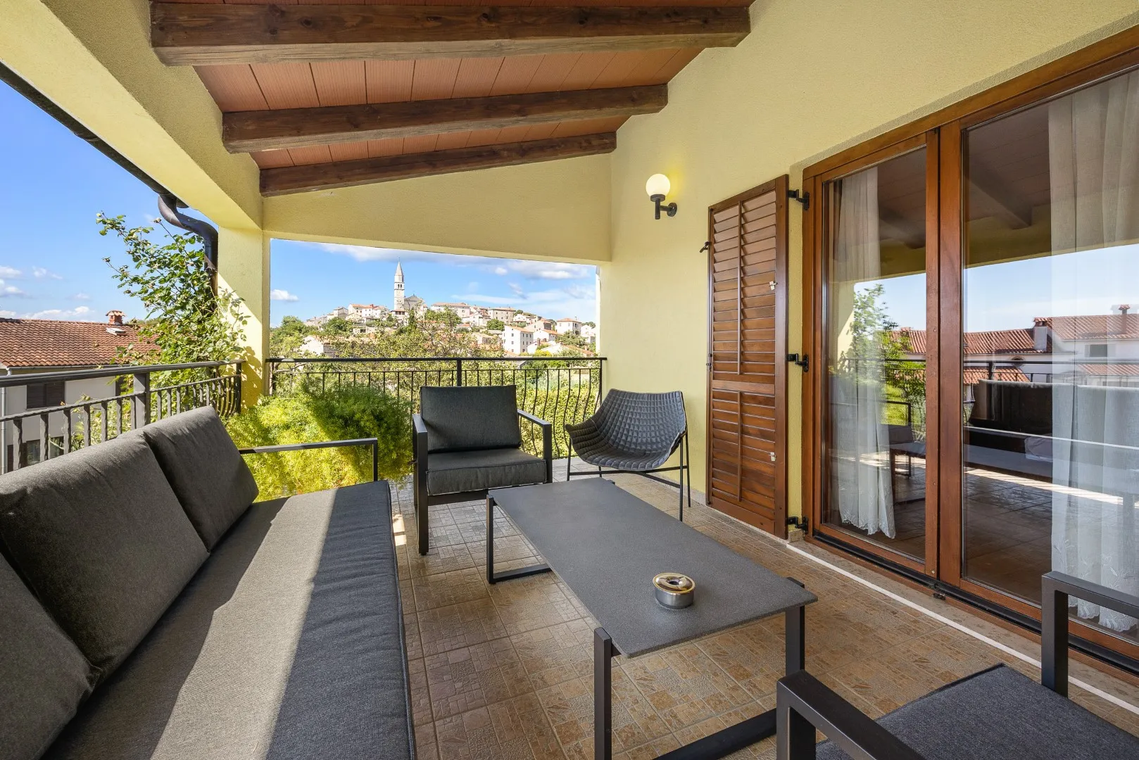 Ein überdachter Balkon mit Loungemöbeln, gegenüber auf dem Höhenrücken der Glockenturm